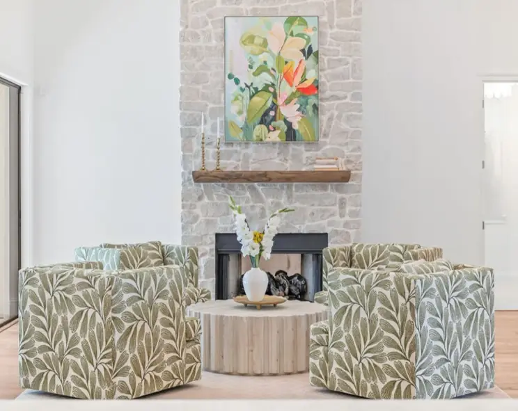 Cozy living room with four green leafy armchairs, a round table with a vase, stone fireplace, framed floral artwork, and light decor.